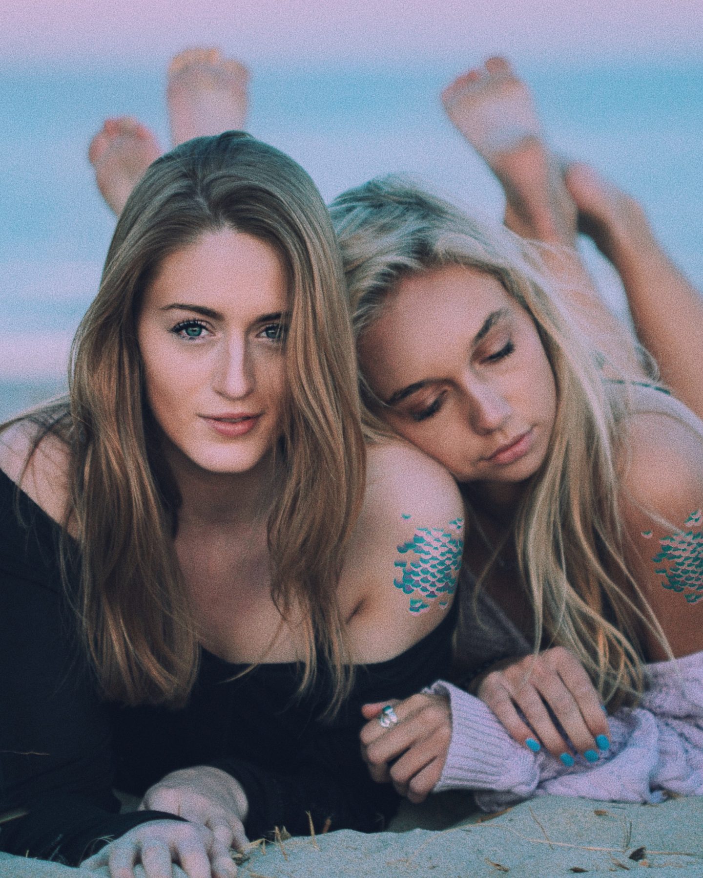 Two women on a beach wearing mermaid scale temporary tattoos.