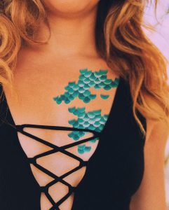Woman in a black criss-cross swimsuit wearing blue temporary tattoos of mermaid scales