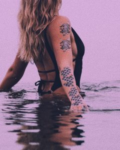 Woman in a black swimsuit wading through water wearing mermaid scale temporary tattoos.